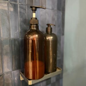 Due flaconi dispenser marroni con pompa su una mensola metallica, appoggiati su una parete di piastrelle blu scuro.