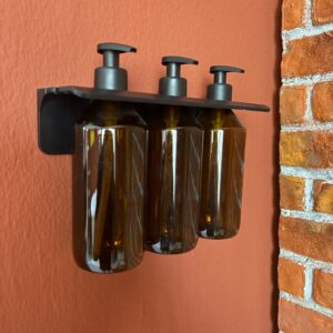 Dispenser a parete nero con tre flaconi ambrati e pompe nere, montato su una parete arancione accanto a un muro di mattoni.