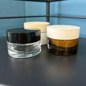 Tre vasetti cosmetici: uno in vetro trasparente con coperchio nero, uno in vetro ambrato con coperchio bianco, su superficie scura.