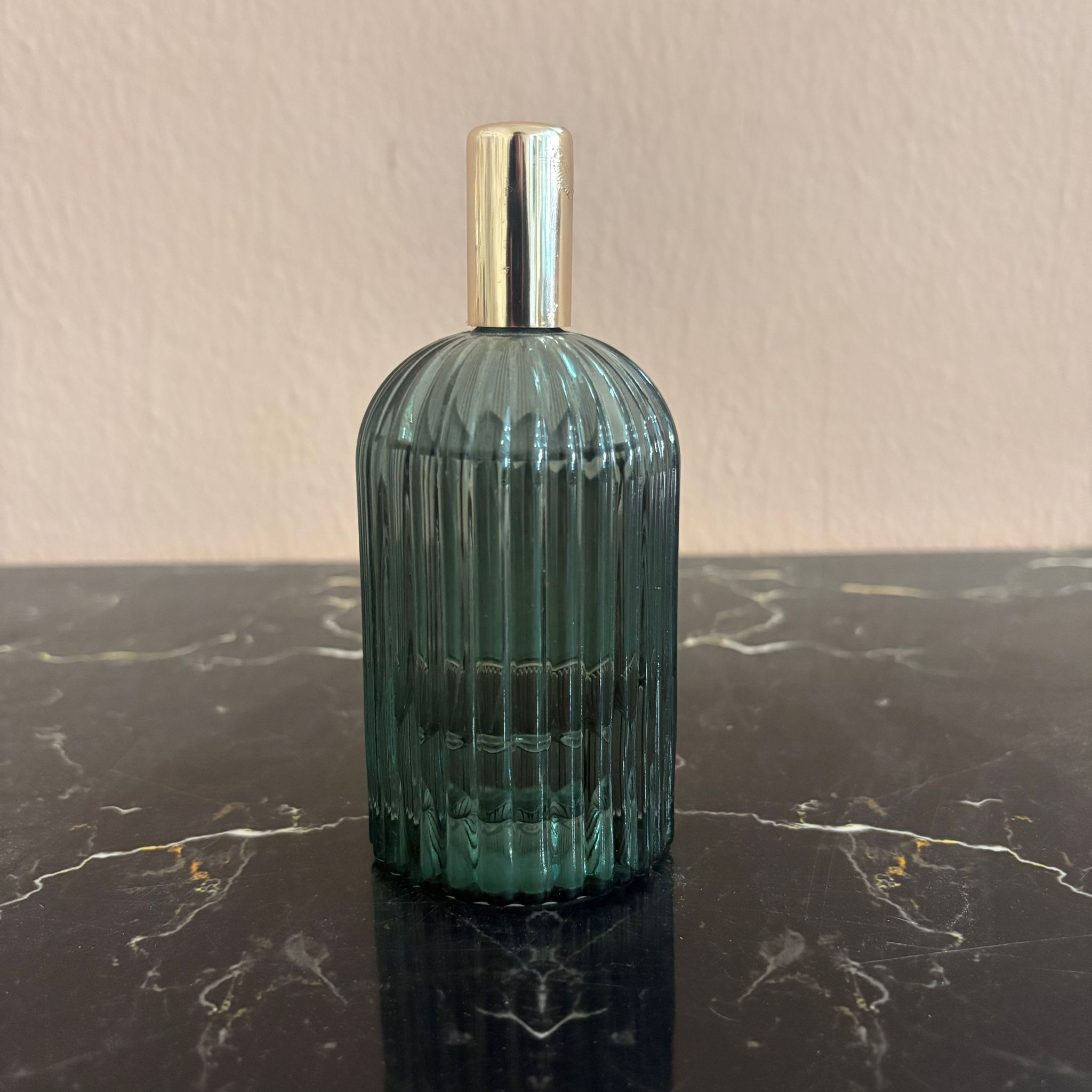 Flacone decorativo in vetro scanalato verde teal con tappo cilindrico dorato, posizionato su una superficie di marmo nero.