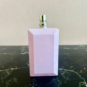 Flacone profumo decorativo rettangolare lilla con motivo floreale in rilievo e tappo erogatore dorato, su superficie scura.