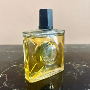 Flacone di profumo vintage in vetro trasparente con liquido ambrato e tappo ottagonale nero, su superficie marmorizzata.