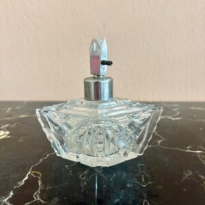 Flacone di profumo vintage in vetro tagliato trasparente con motivi geometrici e atomizzatore metallico argentato su superficie scura.