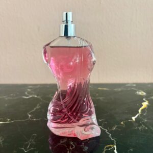 Flacone di profumo in vetro trasparente con design scultoreo e liquido rosa intenso, con vaporizzatore argentato, su superficie scura.