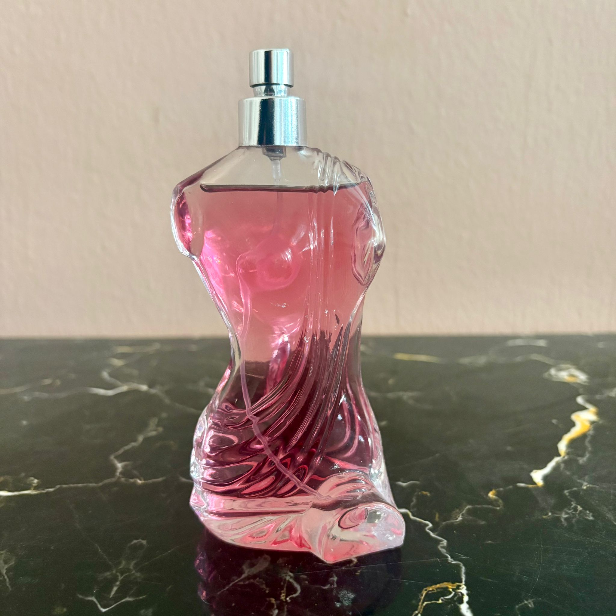 Flacone di profumo in vetro trasparente con design scultoreo e liquido rosa intenso, con vaporizzatore argentato, su superficie scura.