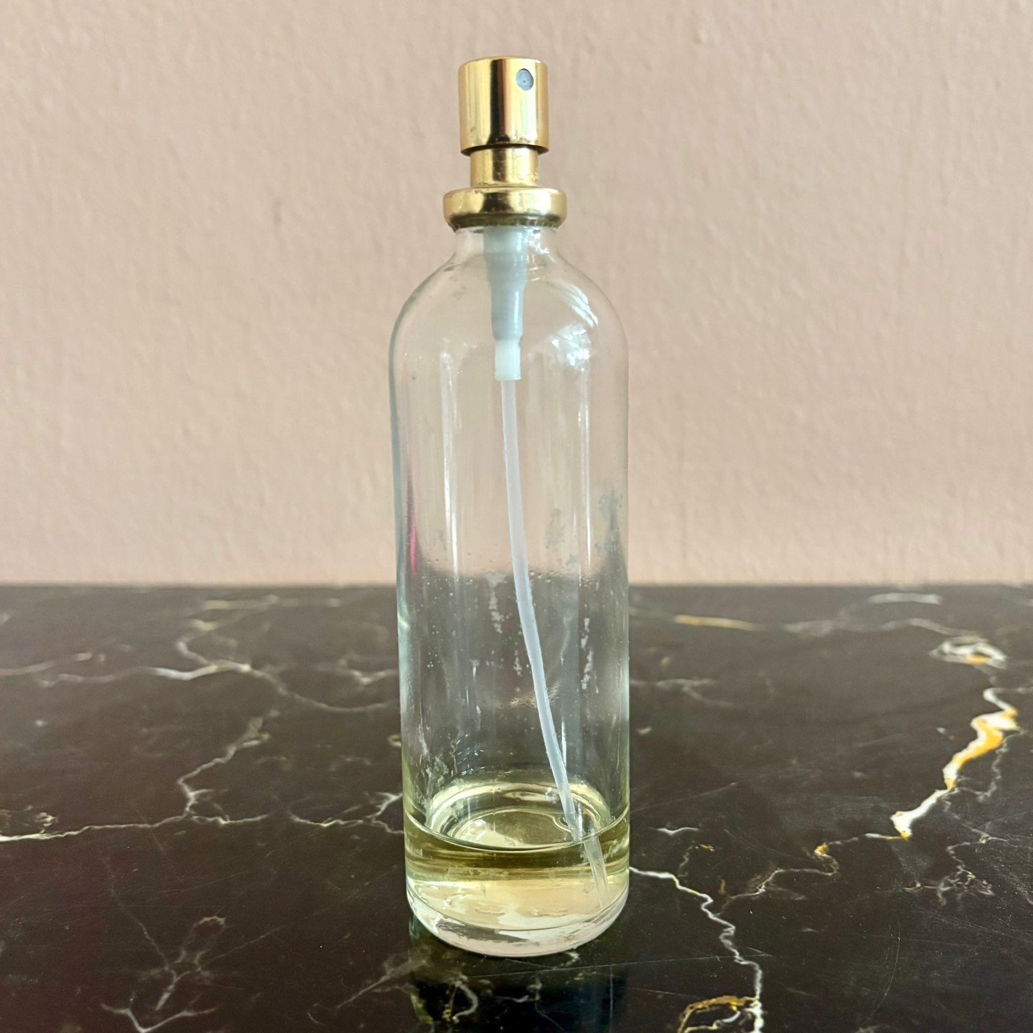 Flacone spray in vetro trasparente con pompa dorata e liquido ambrato sul fondo, posizionato su una superficie di marmo scuro.