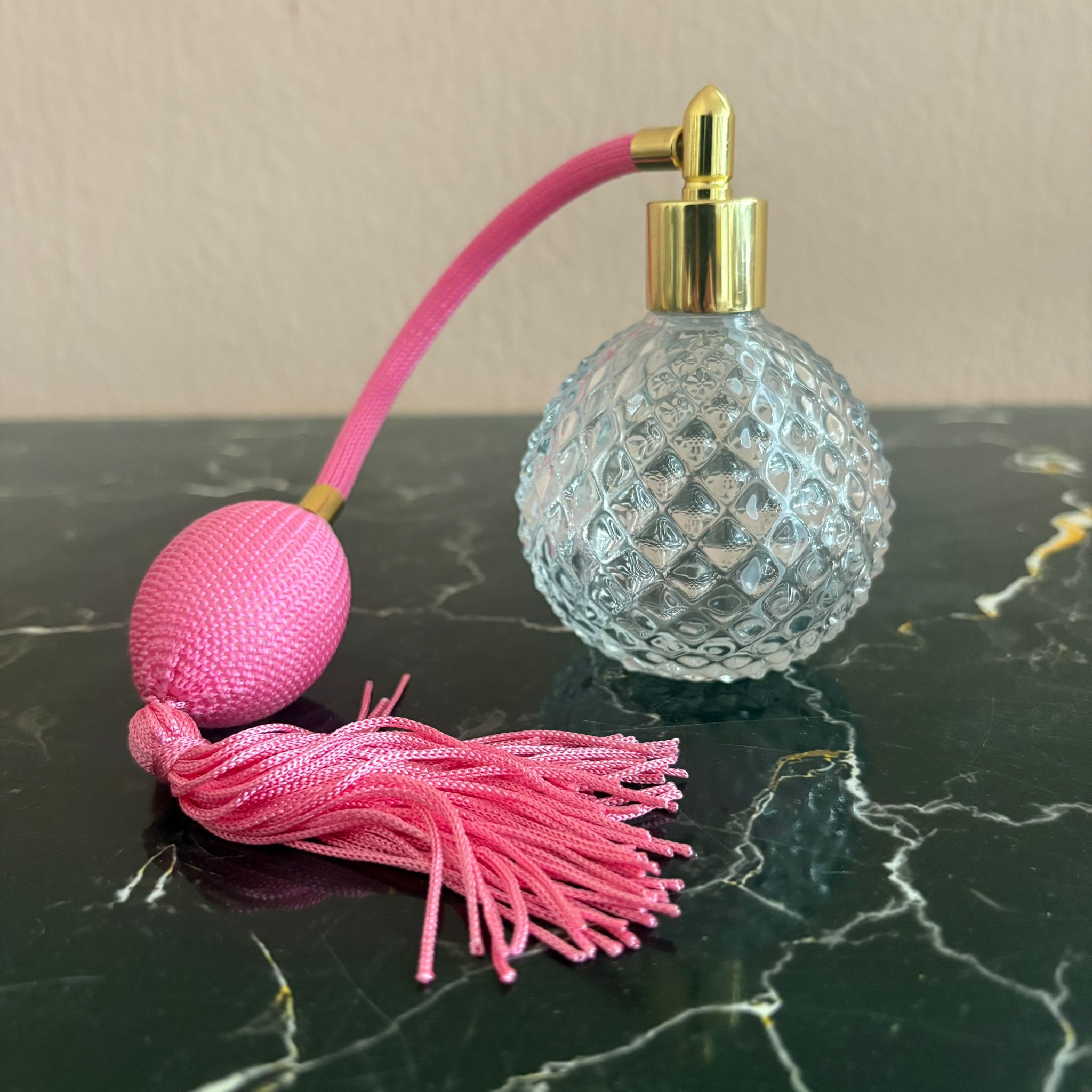 Flacone di profumo vintage in vetro trasparente con motivo a rombi, vaporizzatore dorato e pompetta con nappa rosa su superficie scura.