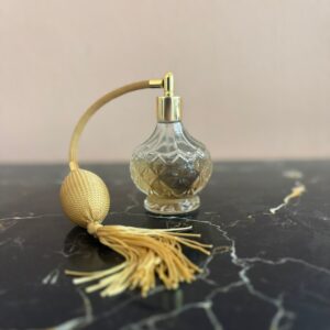 Flacone profumo vintage in vetro con motivo a rombi, atomizzatore dorato a pompetta e nappa, su superficie scura.