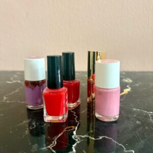 Un set di tre smalti colorati (viola, rosso, rosa) e due rossetti dorati disposti su una superficie di marmo scuro.