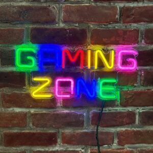 Insegna neon LED multicolore che forma le parole 'GAMING ZONE' su un muro di mattoni rossi, con cavo di alimentazione visibile.