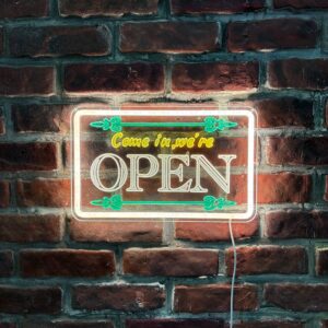 Insegna luminosa a LED con scritta "Come in, we're OPEN" in giallo e bianco, con decorazioni verdi, montata su una parete di mattoni rossi.