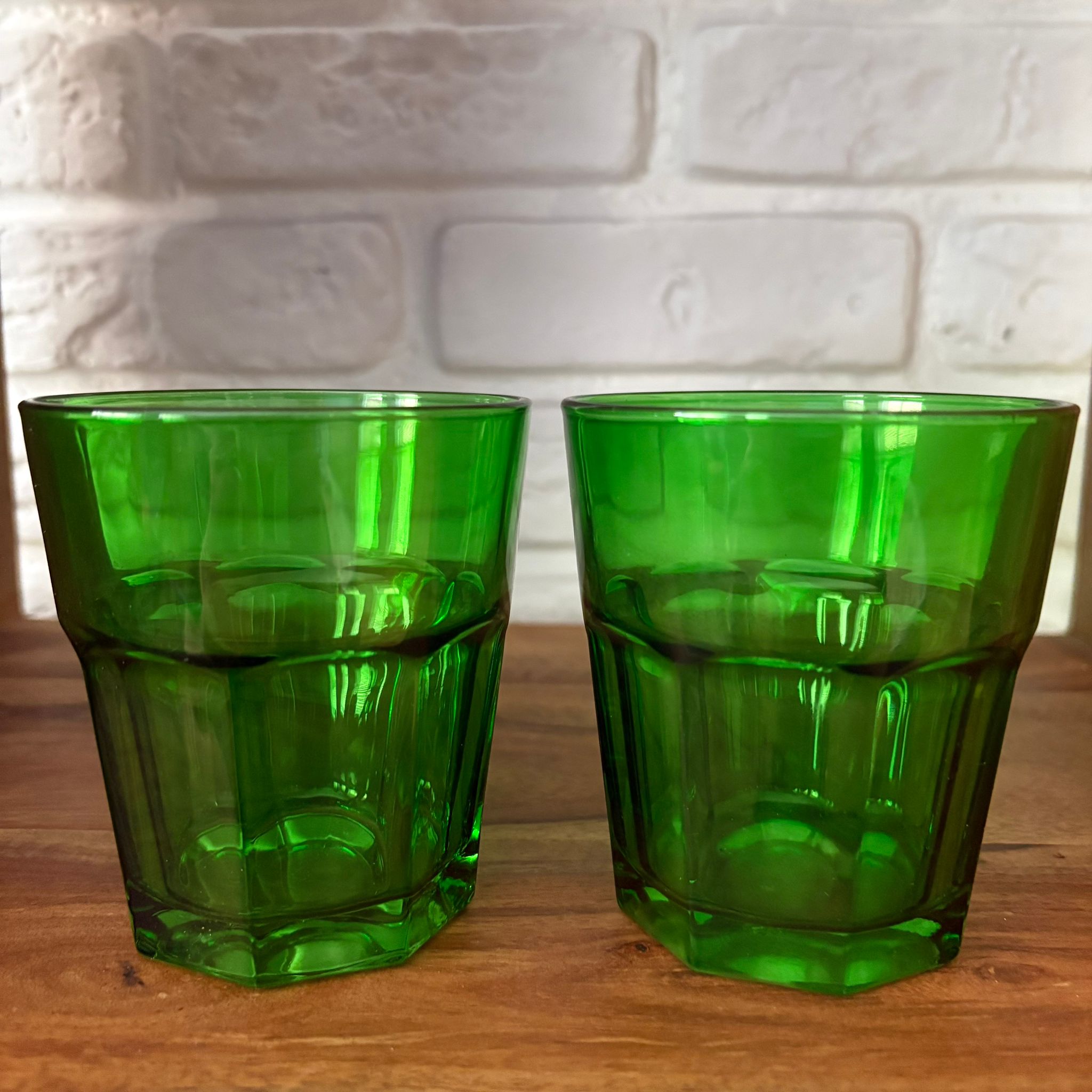 Due bicchieri tumbler in vetro verde intenso, con design sfaccettato e base esagonale, posti su una superficie di legno.