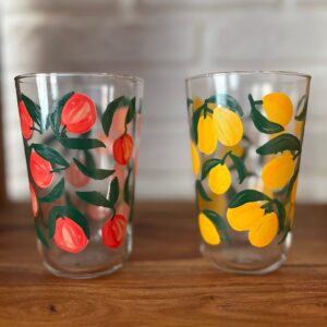 Due bicchieri in vetro trasparente con decorazioni di frutta dipinte: uno con pesche rosse, l'altro con limoni gialli, su superficie di legno.