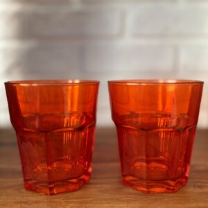 Due bicchieri facettati di colore arancione intenso e traslucido, posizionati su una superficie di legno con sfondo sfocato.