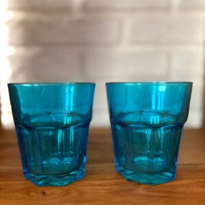 Due bicchieri tumbler in vetro blu sfaccettato, posizionati su una superficie di legno con uno sfondo di mattoni bianchi sfocati.