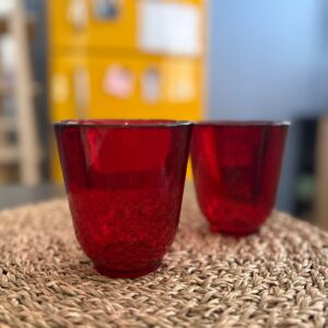 Due bicchieri in vetro rosso con texture a bolle, posizionati su una tovaglietta intrecciata di paglia, con sfondo sfocato.
