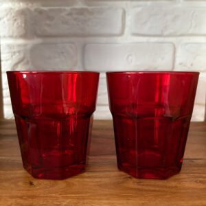 Due bicchieri in vetro rosso intenso con design sfaccettato, posizionati su una superficie di legno davanti a un muro di mattoni bianchi.