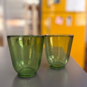 Due bicchieri in vetro verde con superficie a coste verticali, posti su un tavolo grigio con sfondo sfocato giallo.