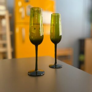 Due calici flute alti in vetro riciclato di colore verde scuro, posizionati su una superficie scura con sfondo giallo sfocato.