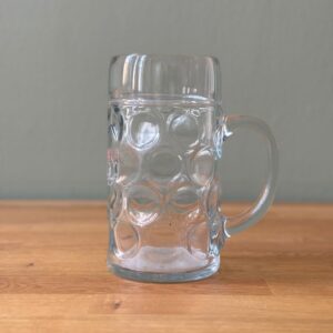 Boccale da birra in vetro trasparente con motivo a fossette e manico robusto, posizionato su un tavolo di legno chiaro.