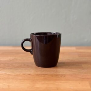 Una tazza in ceramica marrone scuro lucida, con manico, posizionata su un tavolo di legno chiaro con sfondo grigio.