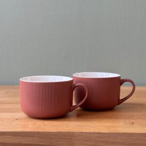 Due tazze in ceramica color terracotta con texture rigata verticale e interno bianco, posizionate su un tavolo di legno.