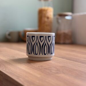 Piccola tazza in ceramica bianca con motivo blu a gocce stilizzate, posizionata su un ripiano in legno chiaro.