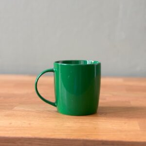 Tazza cilindrica verde brillante con manico, posizionata su un tavolo di legno chiaro con sfondo grigio neutro.