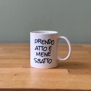 Tazza in ceramica bianca con manico, recante la scritta nera "PRENDO ATTO E MENE SBATTO", posizionata su un tavolo di legno.