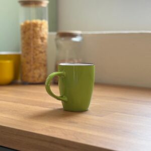 Tazza mug verde brillante in ceramica con bordo nero, posizionata su un piano di lavoro in legno chiaro.