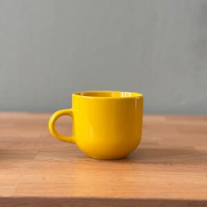 Tazza in ceramica gialla brillante con finitura lucida e manico, posizionata su un tavolo di legno chiaro con sfondo grigio.