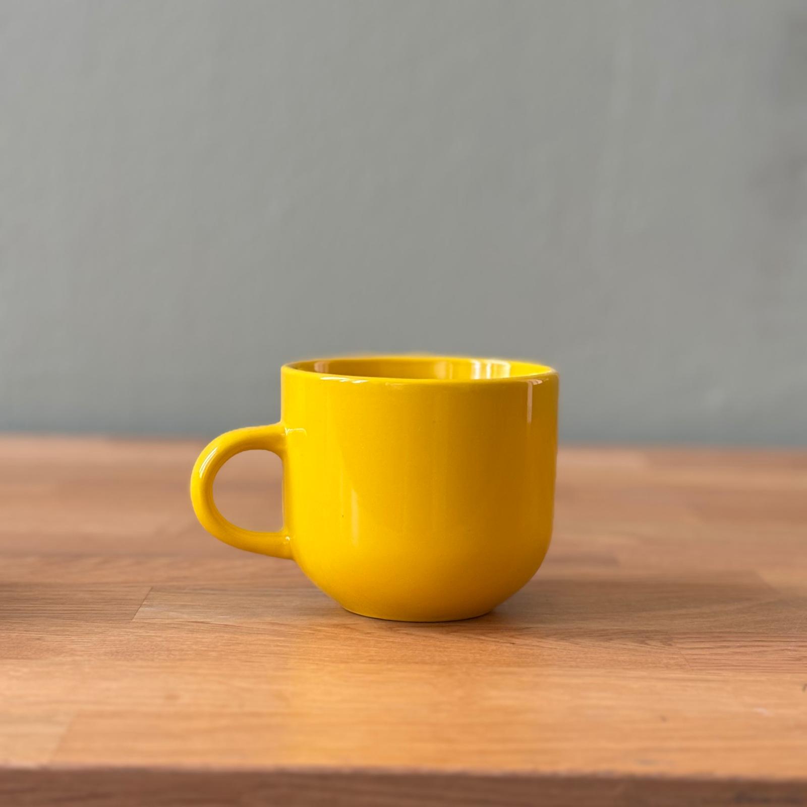 Tazza in ceramica gialla brillante con finitura lucida e manico, posizionata su un tavolo di legno chiaro con sfondo grigio.