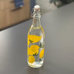 Bottiglia in vetro trasparente con decorazione di limoni gialli e foglie verdi, dotata di tappo ermetico a scatto, su superficie grigia.