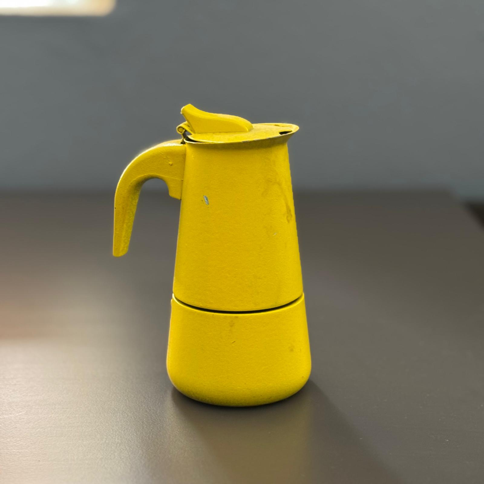 Caffettiera Moka di colore giallo brillante, in metallo verniciato, posizionata su una superficie scura con sfondo grigio.