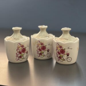 Tre barattoli in ceramica color crema con coperchio, decorati con rose rosse e biciclette, su una superficie scura.