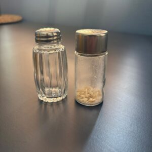Due dispenser in vetro e metallo, una saliera e una pepiera, su una superficie scura, con luce laterale.