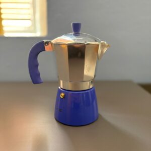 Caffettiera moka bicolore con base blu e corpo superiore argentato, manico e pomello blu, posizionata su un tavolo marrone.