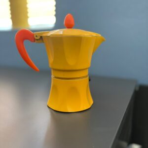 Caffettiera Moka di colore giallo brillante con manico e pomello arancioni, posizionata su una superficie scura.