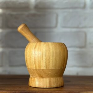 Mortaio e pestello in bambù chiaro su un tavolo di legno scuro, con uno sfondo sfocato di mattoni bianchi.