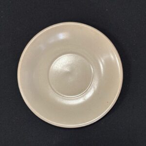 Piattino sottotazza rotondo in ceramica di colore beige chiaro, con bordo leggermente rialzato e incavo centrale, su sfondo scuro.