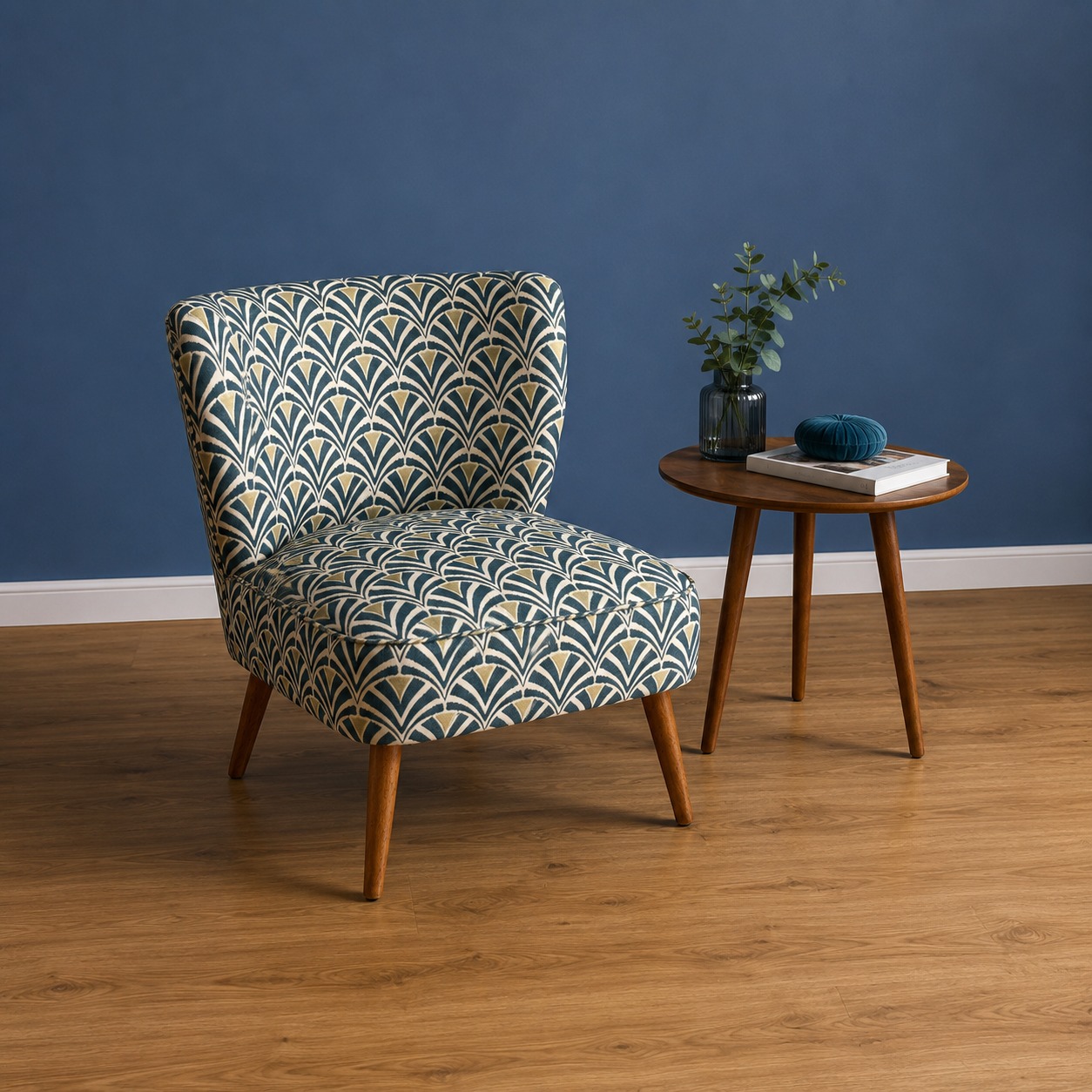 Poltrona accent con rivestimento in tessuto a motivo geometrico Art Déco verde, crema e oro, su gambe in legno, posta su pavimento in parquet davanti a parete blu.