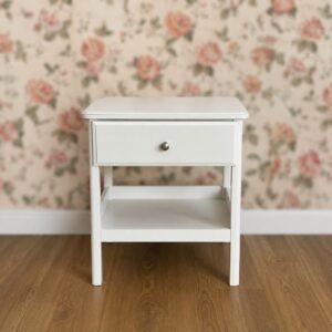 Comodino bianco moderno con un cassetto e un ripiano inferiore, su pavimento in legno, sfondo sfocato con carta da parati floreale.