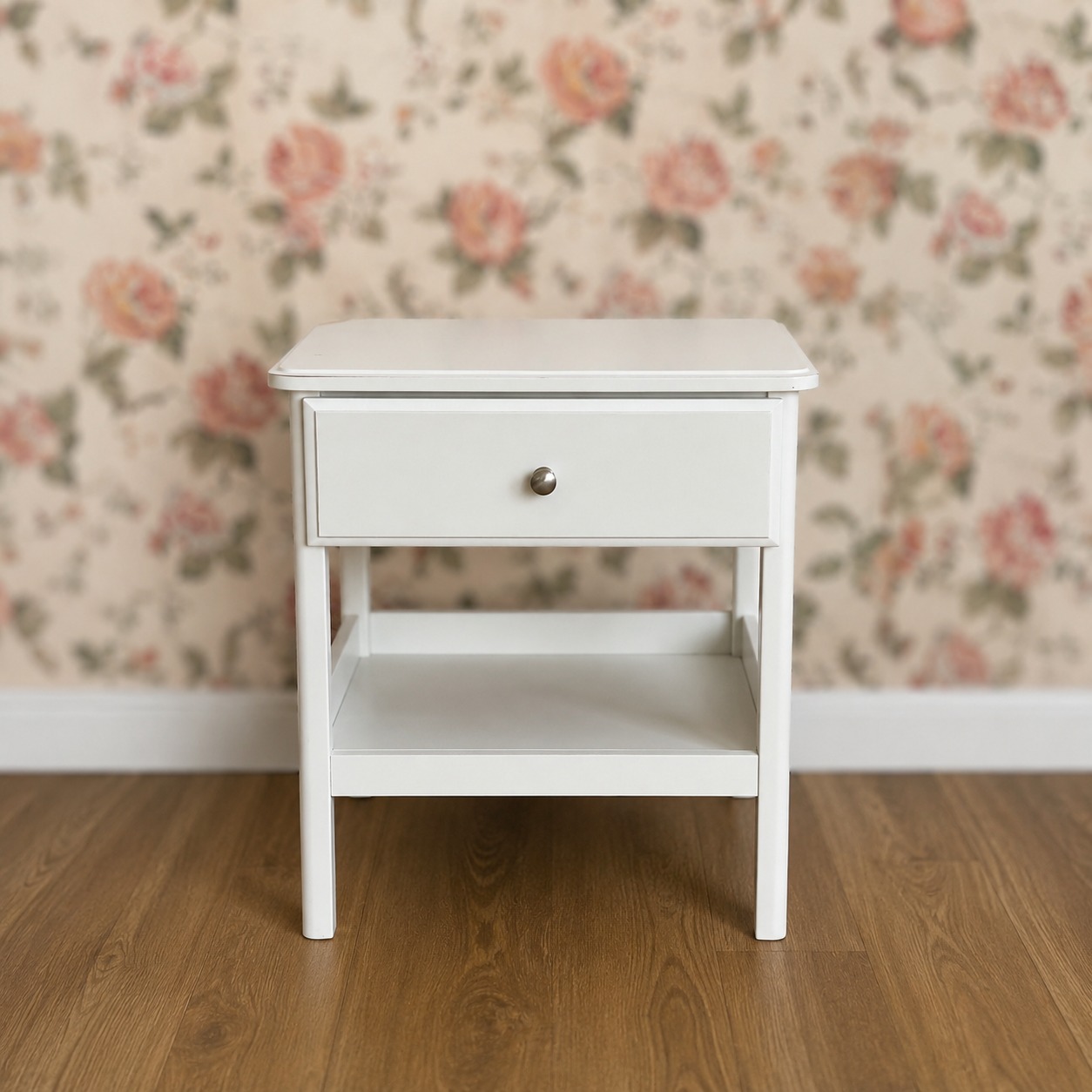 Comodino bianco moderno con un cassetto e un ripiano inferiore, su pavimento in legno, sfondo sfocato con carta da parati floreale.