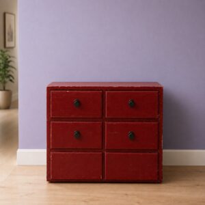 Cassettiera vintage rossa a sei cassetti con finitura usurata e pomelli neri, su pavimento in legno chiaro davanti a parete lilla.