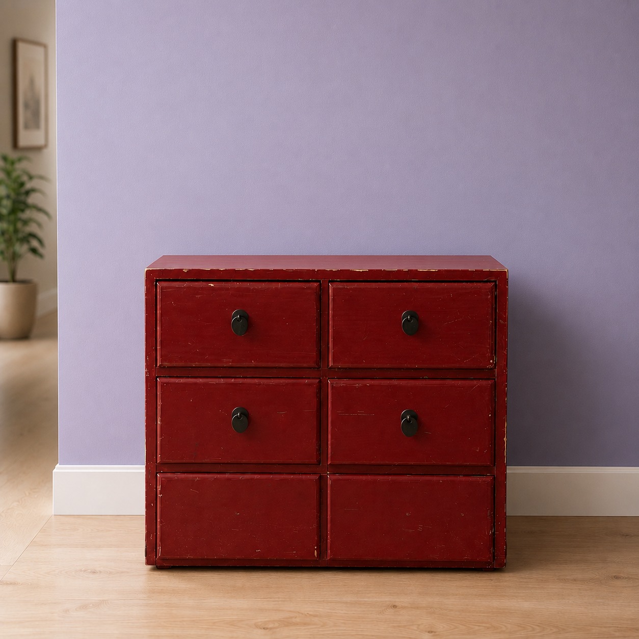 Cassettiera vintage rossa a sei cassetti con finitura usurata e pomelli neri, su pavimento in legno chiaro davanti a parete lilla.
