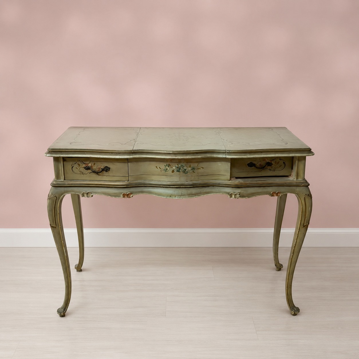 Consolle toeletta antica in legno laccato verde salvia con decorazioni floreali e gambe cabriole, su sfondo rosa.