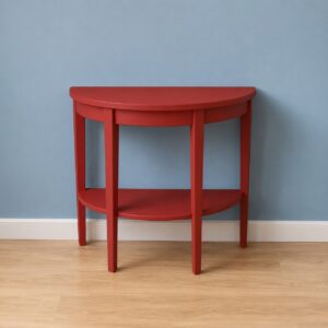 Consolle semicircolare rossa con ripiano inferiore, appoggiata a una parete blu chiaro su pavimento in legno.