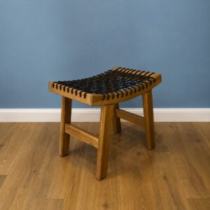 Sgabello in legno chiaro con seduta intrecciata di corda nera, posizionato su un pavimento in parquet davanti a una parete blu.