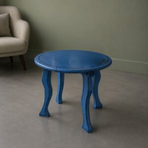 Tavolino rotondo blu con gambe curve e finitura anticata, posizionato su pavimento grigio chiaro accanto a una poltrona beige.