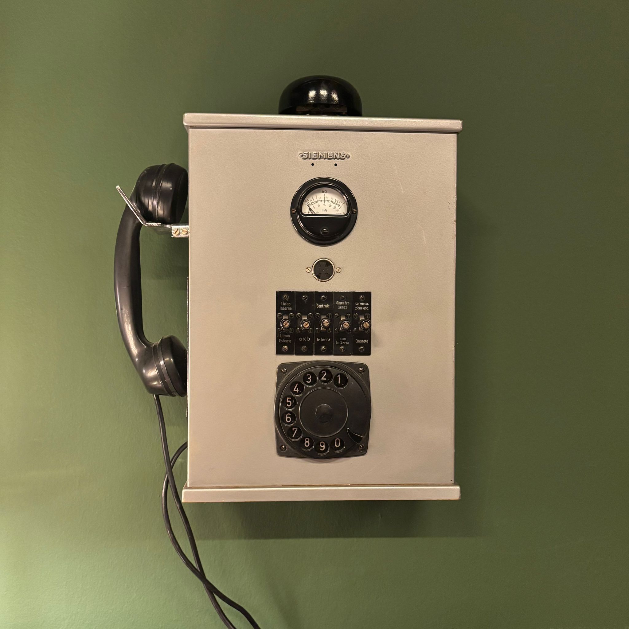 Telefono da parete Siemens vintage grigio con cornetta nera, quadrante rotante, misuratore analogico e interruttori a levetta, su sfondo verde.
